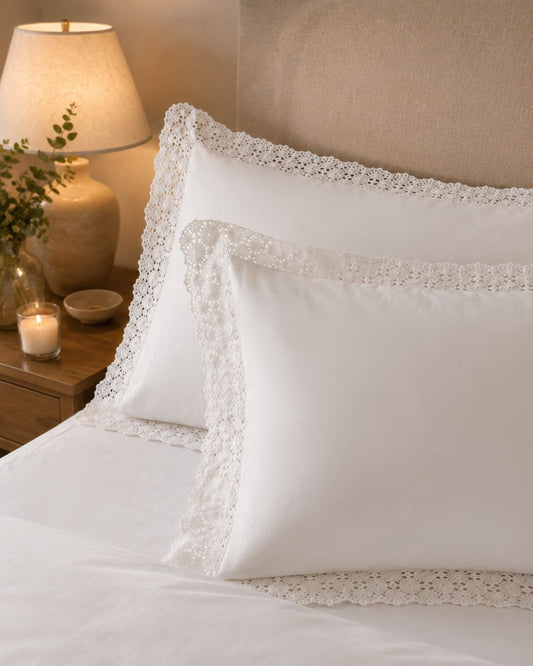 White decorative pillows with intricate white lace trim, styled on a bed with a beige upholstered headboard, a lit bedside lamp, and a candle.