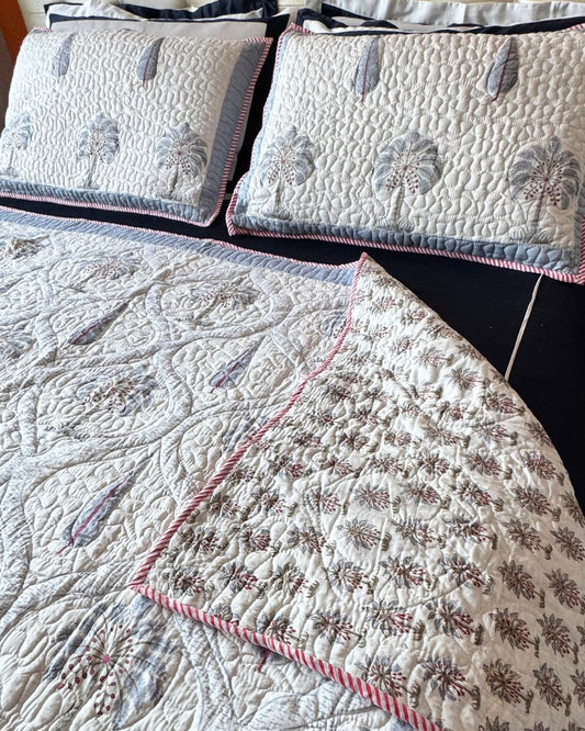 A quilted bedding set featuring a cream-colored quilt and matching pillowcases with embroidered palm tree motifs, pink and blue-gray accents, and pink striped piping.
