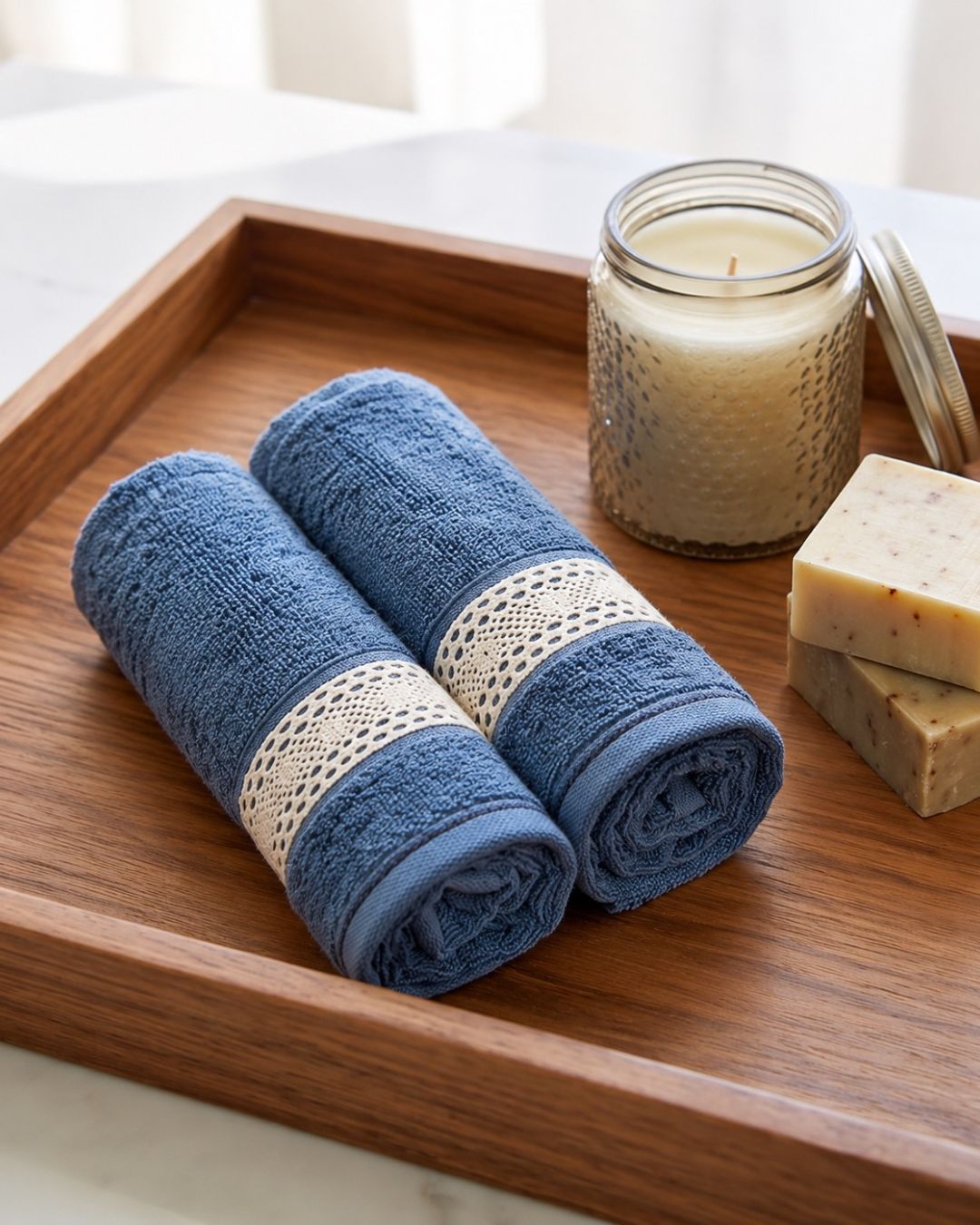 Two rolled blue bath towels with cream lace bands are arranged on a wooden tray beside a candle and soap bars in a bright bathroom setting.