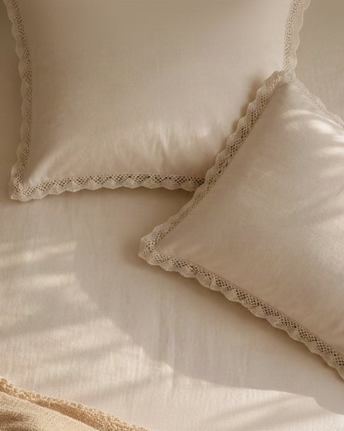 Two cream-colored decorative pillows with scalloped crochet lace trim, styled on a matching cream bedspread with soft sunlight shadows and a crocheted throw blanket in the corner.