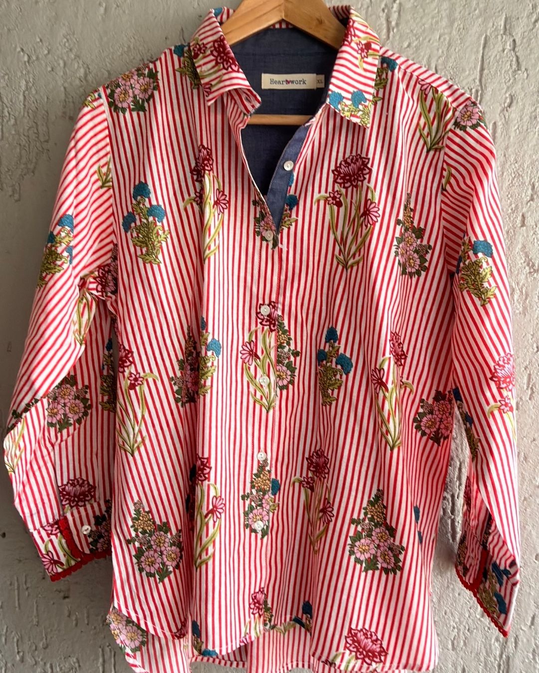 A long-sleeve button-up shirt by  featuring red and white vertical stripes with a colourful floral pattern, hanging on a wooden hanger 
