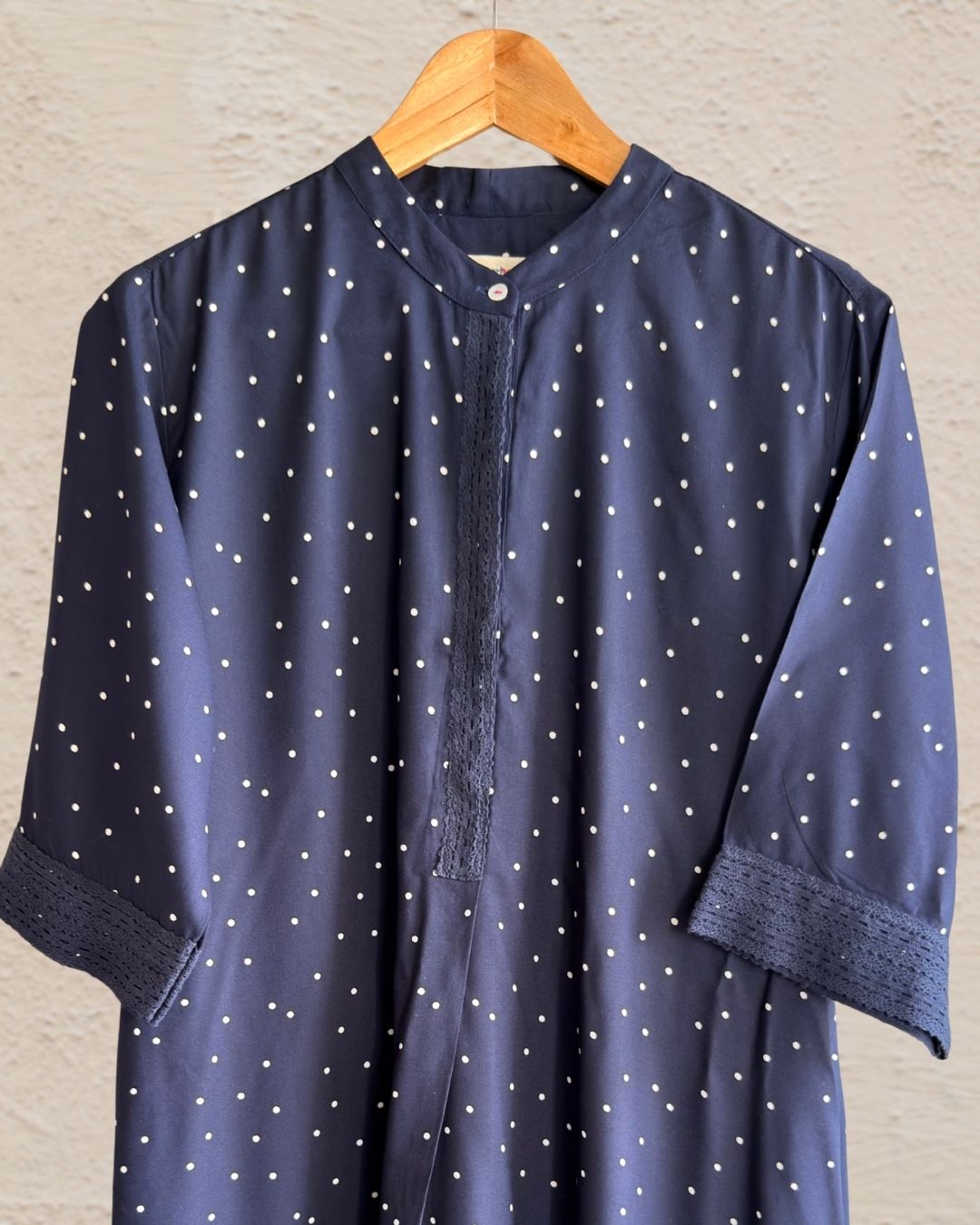 A navy blue cotton tunic with a white polka dot pattern, hanging on a wooden hanger against a textured beige wall.