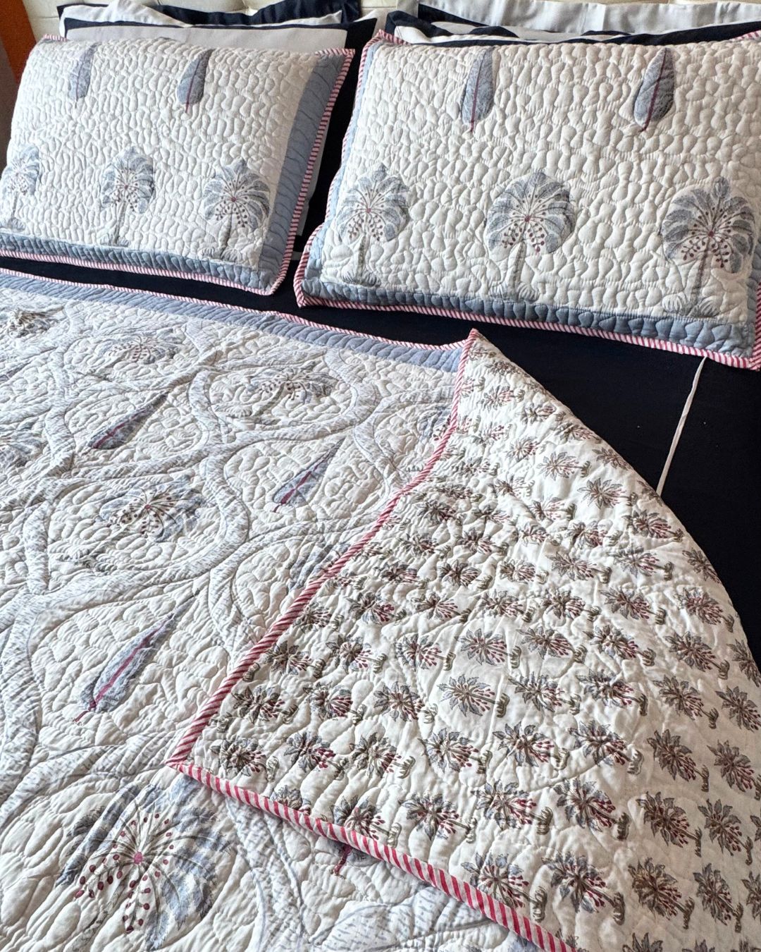 A quilted bedding set featuring a cream-colored quilt and matching pillowcases with embroidered palm tree motifs, pink and blue-gray accents, and pink striped piping.