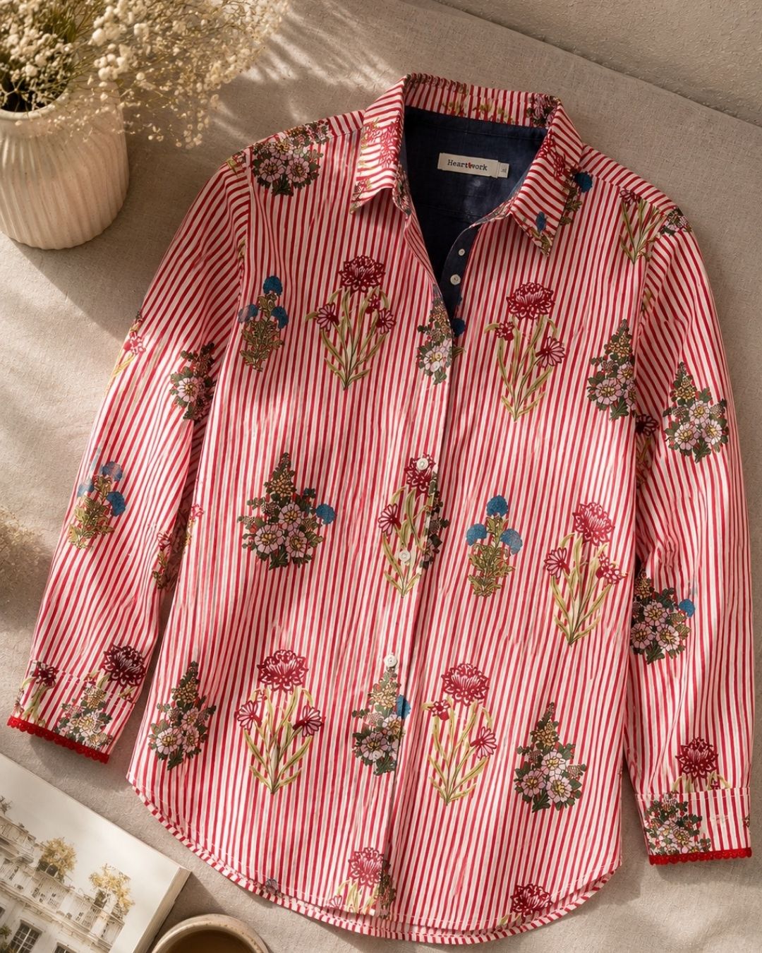 A long-sleeve button-up shirt with red and white vertical stripes and embroidered floral motifs, laid flat on a beige textured surface with a small dried flower arrangement in the background.