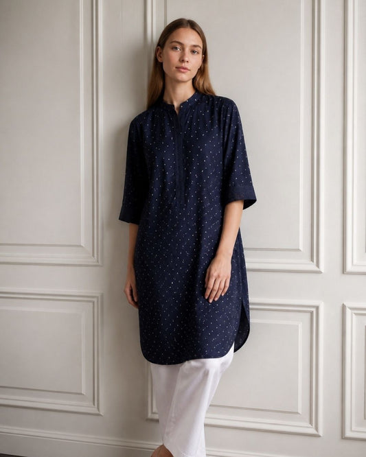 A woman stands against a white paneled wall wearing a navy blue tunic top with tiny white polka dots and white pants.